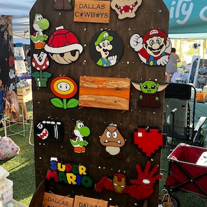 Puede incluir: Un tablero de madera con varios recortes de madera pintados a mano de personajes populares de videojuegos, incluyendo Mario, Luigi, Yoshi y un Baby Yoda. El tablero también presenta algunos letreros de madera con el texto "Dallas Cowboys".