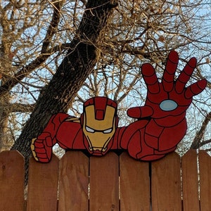 Könnte beinhalten: Eine rote und gelbe Iron-Man-Figur, die über einen Holzzaun lugt. Die Hand der Figur ist in einer Superheldenpose erhoben.