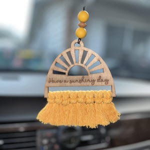 May include: Wooden car air freshener with a sunburst design and yellow macrame fringe. The text "Have a sunshining day" is engraved on the wooden piece. The air freshener is hanging from a black string with yellow and brown beads.