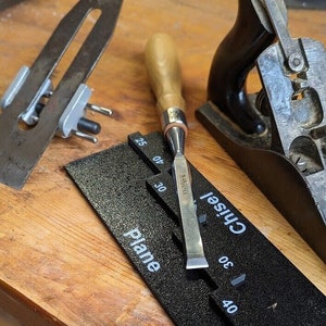 Op de afbeelding: Een verzameling houtbewerkingsgereedschappen, waaronder een beitel, een schaaf en een slijphulp voor messen. De beitel heeft een houten handvat en een metalen lemmet. De slijphulp is zwart met witte markeringen. De schaaf is zwart en zilver.