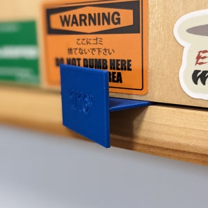 May include: A blue 1/16" corner bracket is attached to a wooden shelf. The bracket is positioned to support a cardboard box with a warning label in English and Japanese. The label reads "WARNING" and "DO NOT DUMB HERE AREA."