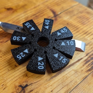 Puede incluir: Un calibrador de ángulos negro con marcas blancas, incluyendo números y flechas, descansa sobre una superficie de madera. El calibrador se utiliza con una herramienta de metal, probablemente para carpintería o metalurgia. Los números indican ángulos de 10 a 45 grados.