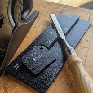 May include: A woodworking chisel with a wooden handle and a metal blade rests on a black shim with measurements. A metal plane is in the background. The shim has the text "1/8 Shim" and measurement markings.