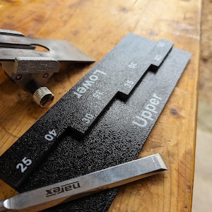 Puede incluir: Primer plano de herramientas de carpintería sobre una superficie de madera. Las herramientas incluyen un calibrador negro texturizado con marcas blancas, un cincel plateado con la marca "narex" y una herramienta de metal con un tornillo.