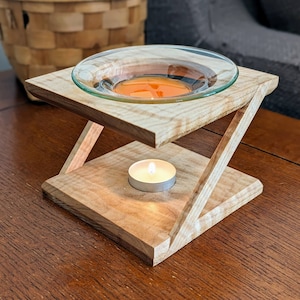 Puede incluir: Un difusor de aceites esenciales de madera con un plato de vidrio transparente que contiene líquido naranja. Debajo hay una vela de té encendida. El marco de madera tiene un acabado natural.