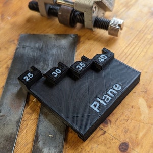 May include: A metal plane blade sharpening jig with a black 3D-printed angle guide. The guide has the word "Plane" and numbers 25, 30, 35, and 40 printed on it. The jig is on a wooden surface.