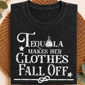 May include: A black crew neck t-shirt with a vintage wash finish. The front features white screen-printed text that reads "TEQUILA MAKES HER CLOTHES FALL OFF" with a small bottle icon replacing the letter Q and two star accents at the bottom.