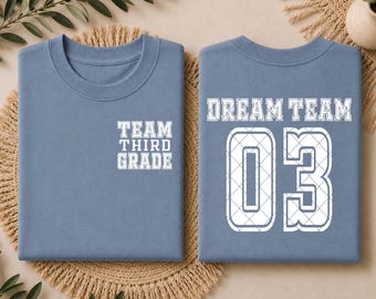 May include: A blue t-shirt featuring white text on the front left chest that reads TEAM THIRD GRADE. The back of the shirt displays the text DREAM TEAM above the large number 03, both in white with a grid pattern detail on the lettering and numbers.