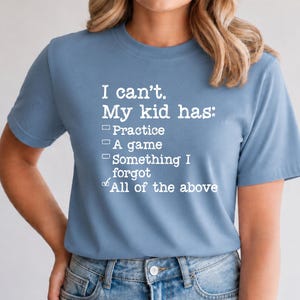 May include: A light blue t-shirt with the text "I can't. My kid has: Practice, A game, Something I forgot, All of the above" in white. The shirt is made of a soft material and has short sleeves.