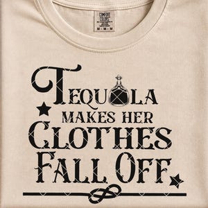 Op de afbeelding: Beige t-shirt met de zwarte tekst "Tequila Makes Her Clothes Fall Off". Het ontwerp bevat een tequila fles afbeelding, sterren en decoratieve elementen. Het shirt is van het merk Comfort Colors.