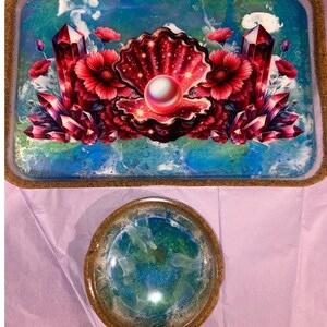 May include: A rectangular tray and a circular dish with a cork-like rim. The tray features a central open seashell containing a white pearl, flanked by red flowers and dark pink crystals against a blue and white swirling background.
