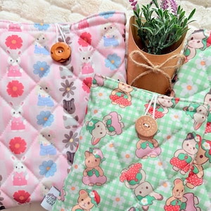 May include: Quilted fabric pouches in pink and green, featuring repeating patterns of bunnies, bears, and flowers. Each pouch has a wooden button closure and a diamond-stitched pattern. A small potted plant with purple flowers is also visible.