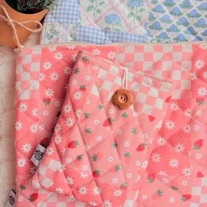 May include: Pink quilted fabric pouches with a strawberry and daisy pattern, and a checkerboard design. One pouch has a wooden button closure. The background features a blue floral and gingham pattern.