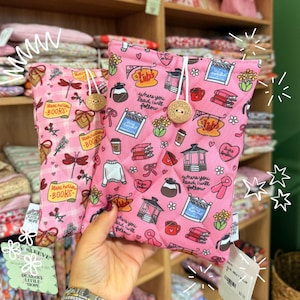 May include: Two pink quilted book sleeves with various patterns, including dragonflies, books, coffee cups, and the text "Stars hollow BOOKS" and "where you lead, I will follow". Each sleeve has a wooden button closure.