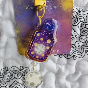May include: A yellow keychain with a clear charm featuring a purple bottle with a moon face and stars. The charm is attached to a smaller clear charm with a moon face. The keychain is labeled "Moon Jar Spirit Art".