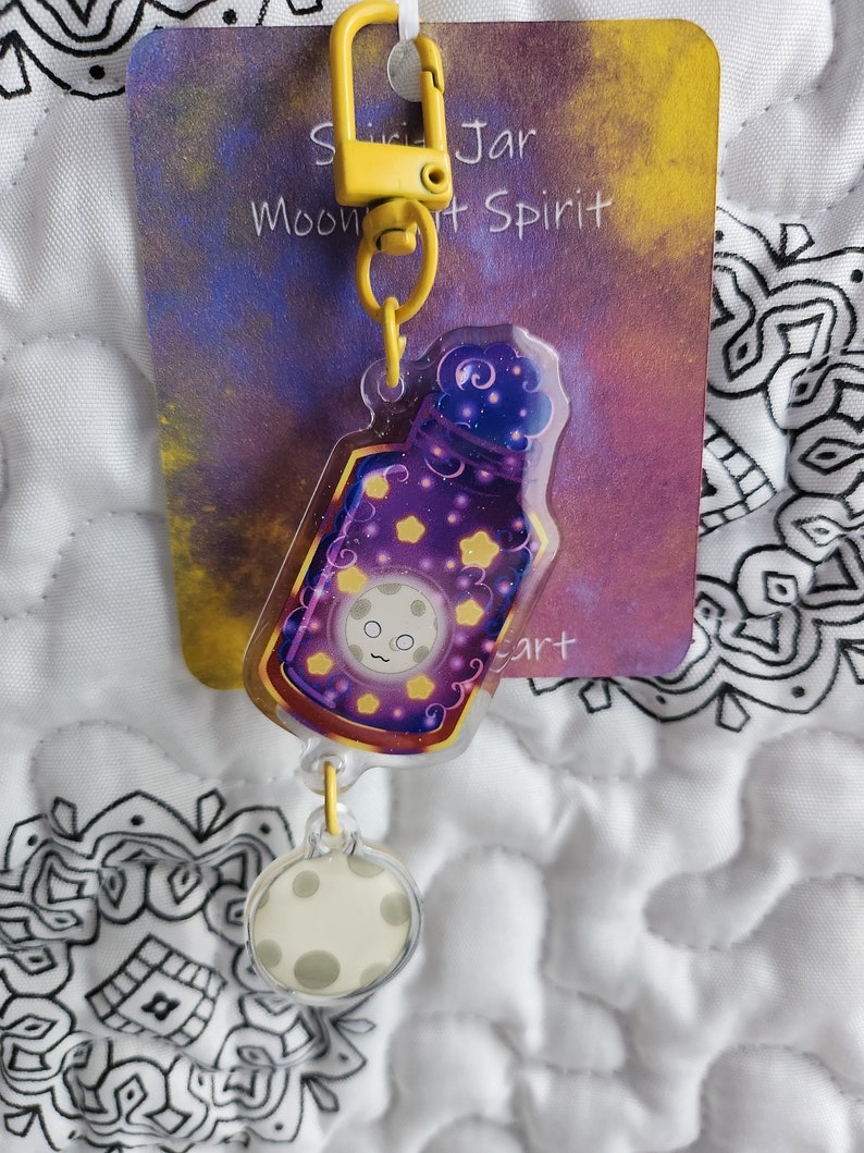 May include: A yellow keychain with a clear charm featuring a purple bottle with a moon face and stars. The charm is attached to a smaller clear charm with a moon face. The keychain is labeled "Moon Jar Spirit Art".