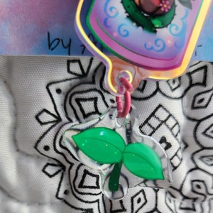 May include: A green leaf charm with a clear plastic background. The charm is attached to a pink and white string.