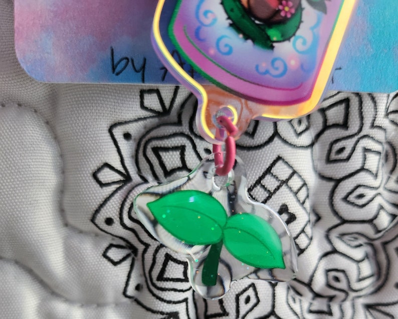 May include: A green leaf charm with a clear plastic background. The charm is attached to a pink and white string.