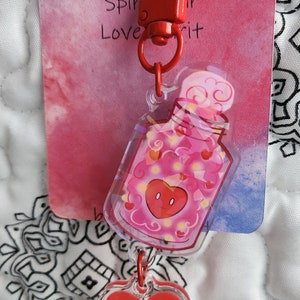 May include: A red and pink heart-shaped charm with a clear plastic bottle filled with pink hearts and a red heart with a face. The bottle is attached to a red metal clasp with a ring. The charm is hanging from a white background with the text "Spir your Love it" on a red and pink background.