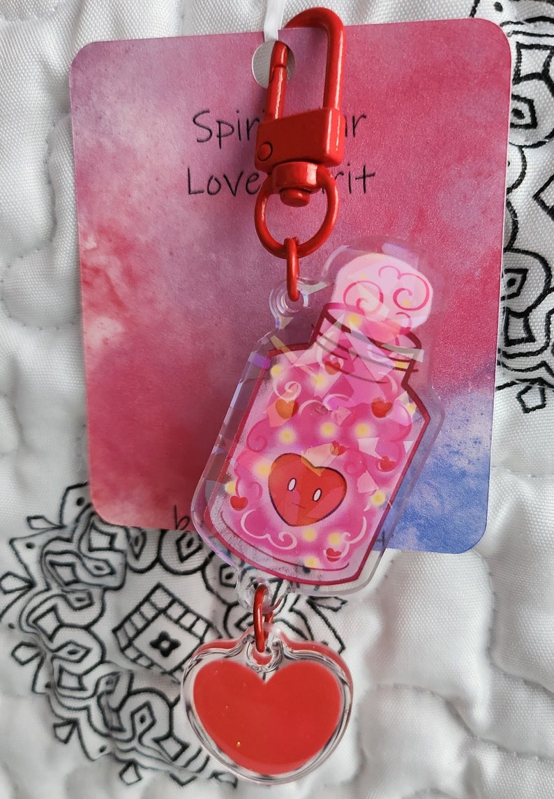 May include: A red and pink heart-shaped charm with a clear plastic bottle filled with pink hearts and a red heart with a face. The bottle is attached to a red metal clasp with a ring. The charm is hanging from a white background with the text "Spir your Love it" on a red and pink background.