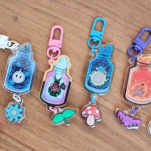 May include: Seven colorful keychains with clear glass bottles filled with glitter and small charms. Each bottle has a different character inside and a charm hanging below. The charms include a heart, snowflake, leaf, mushroom, bat, and moon.