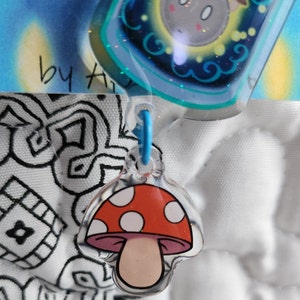May include: A red and white mushroom charm with white dots on the cap and a pink stem. The charm is attached to a blue string.