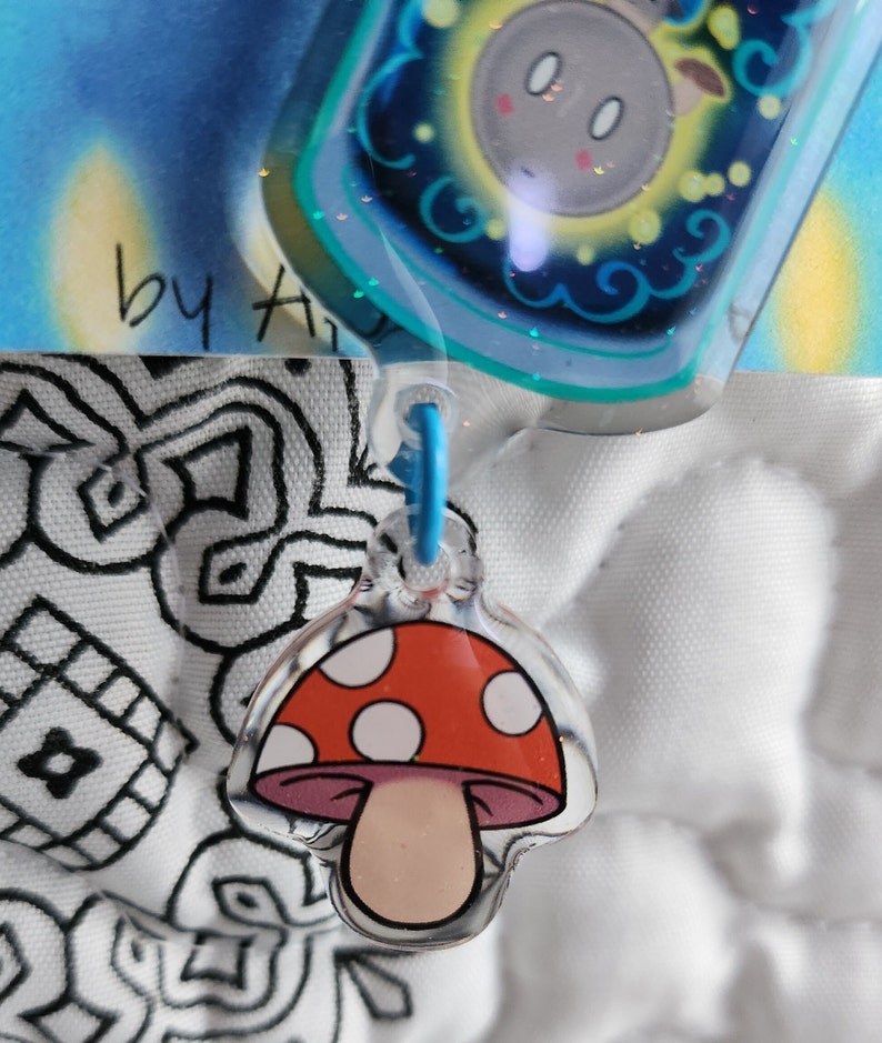 May include: A red and white mushroom charm with white dots on the cap and a pink stem. The charm is attached to a blue string.