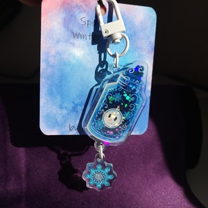 May include: A blue and white winter-themed keychain with a snowman inside a bottle charm and a snowflake charm. The keychain is attached to a white clip.