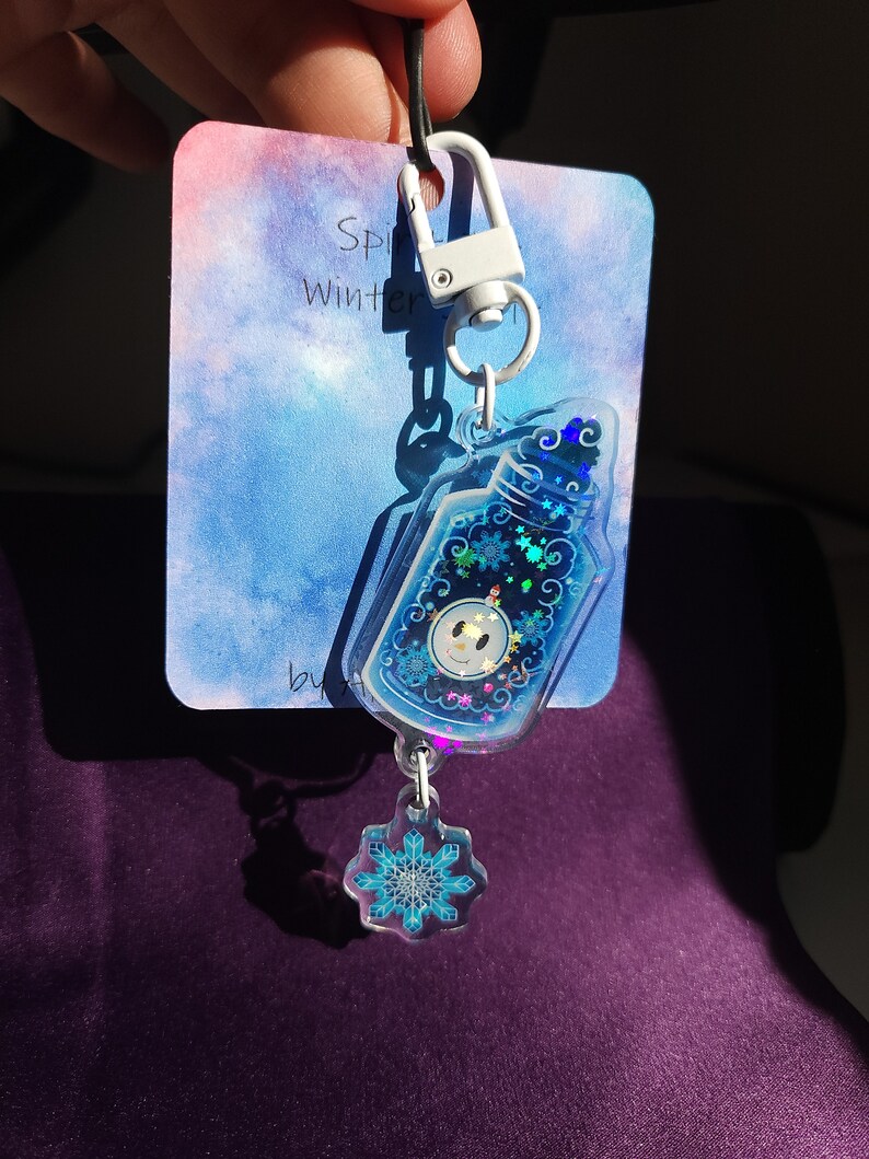 May include: A blue and white winter-themed keychain with a snowman inside a bottle charm and a snowflake charm. The keychain is attached to a white clip.