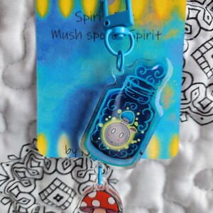 May include: A blue and yellow keychain with a clear charm featuring a mushroom in a bottle. The charm is attached to a blue metal clasp. The keychain has the text "Spirit Mush Spirit" on the card.