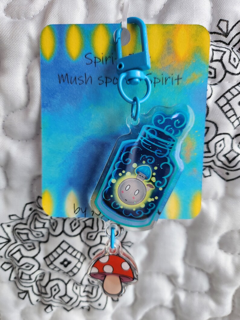 May include: A blue and yellow keychain with a clear charm featuring a mushroom in a bottle. The charm is attached to a blue metal clasp. The keychain has the text "Spirit Mush Spirit" on the card.