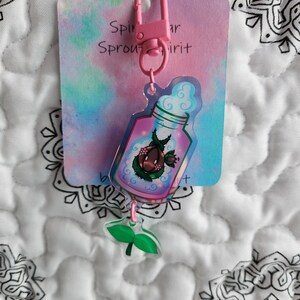 May include: Pink and green keychain with a clear bottle charm containing a small plant and a pink cloud. The charm is attached to a pink metal ring and a card that says "Sprout Spirit".