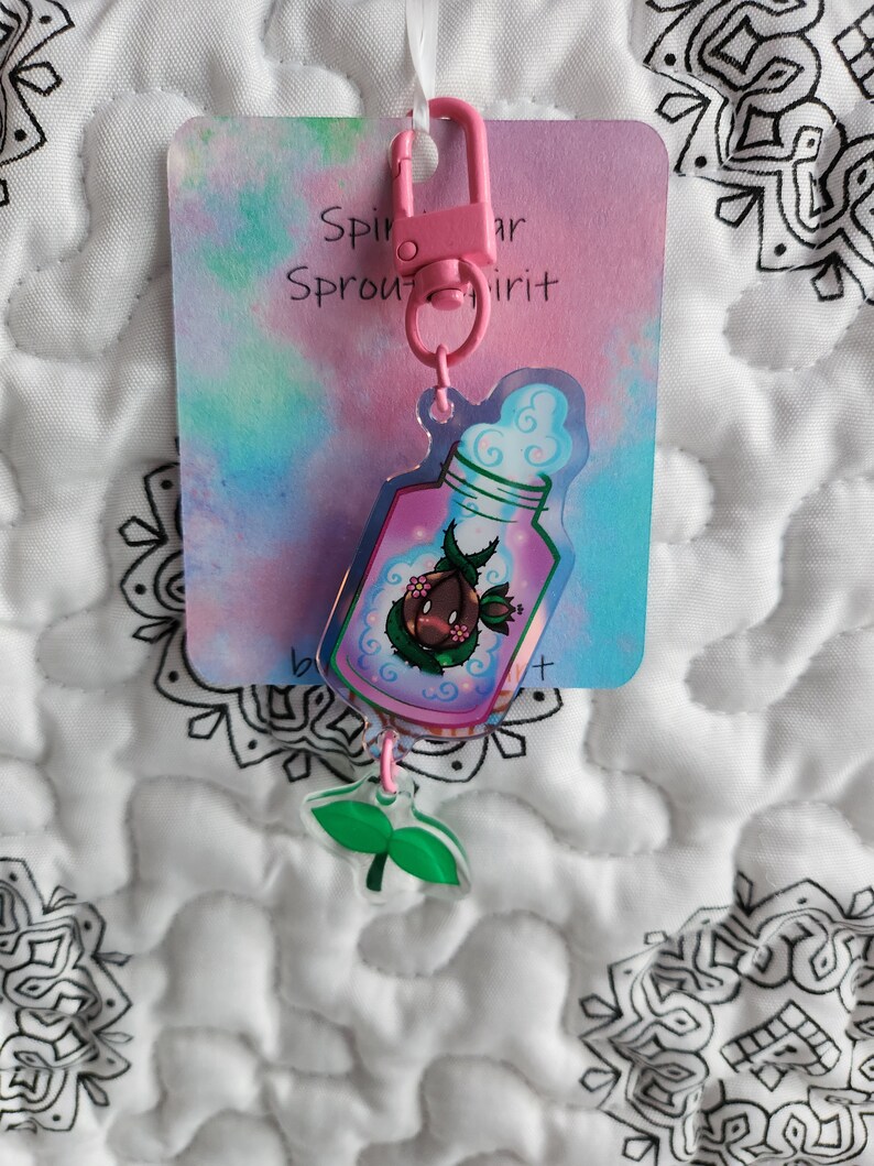 May include: Pink and green keychain with a clear bottle charm containing a small plant and a pink cloud. The charm is attached to a pink metal ring and a card that says "Sprout Spirit".