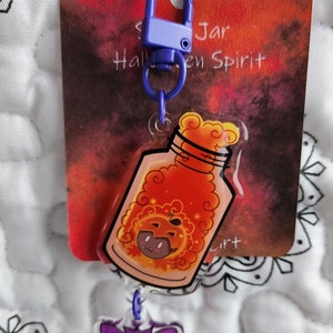 May include: A purple and blue Halloween-themed keychain with a clear jar containing a pumpkin-shaped spirit. The jar has a swirling orange and yellow liquid inside. The keychain also has a purple bat charm.