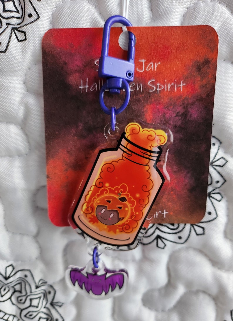 May include: A purple and blue Halloween-themed keychain with a clear jar containing a pumpkin-shaped spirit. The jar has a swirling orange and yellow liquid inside. The keychain also has a purple bat charm.