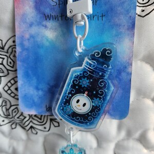 May include: A blue and white winter spirit charm with a snowman inside a bottle and a snowflake charm attached to a white metal key chain. The charm is on a white card with the text "Winter Spirit" on it.
