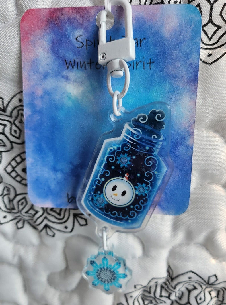 May include: A blue and white winter spirit charm with a snowman inside a bottle and a snowflake charm attached to a white metal key chain. The charm is on a white card with the text "Winter Spirit" on it.