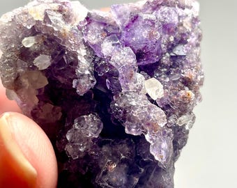 Puede incluir: Primer plano de un grupo de cristales de fluorita en bruto. El cristal presenta un tono púrpura intenso con áreas más claras y translúcidas y pequeños cristales de cuarzo transparente. La textura rugosa y la formación natural son visibles.