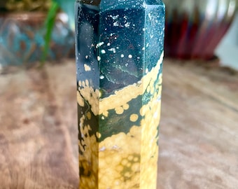 May include: A polished gemstone obelisk with a hexagonal shape. The top is a deep teal color with white speckles, transitioning to a golden-yellow base with green and brown spots. The obelisk is displayed on a wooden surface.