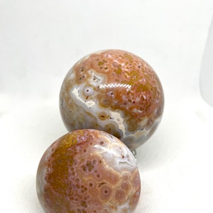 May include: Two Ocean Jasper spheres. The spheres have a mix of colors, including shades of brown, orange, and white. The spheres have a polished, smooth surface and are of different sizes. The larger sphere is positioned behind the smaller one.