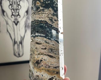 Puede incluir: Un obelisco de piedra preciosa pulida con una parte superior puntiaguda. La piedra presenta una mezcla de colores, incluyendo verde oscuro, beige y marrón, con un patrón veteado. El obelisco se sostiene en una mano, mostrando su tamaño y forma. Un marco artístico está en el fondo.