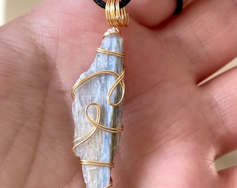May include: A blue kyanite crystal pendant wrapped in gold-colored wire, suspended from a black cord. The crystal is a light blue color with a rough, natural texture. The wire is intricately wrapped around the stone, creating a decorative design.