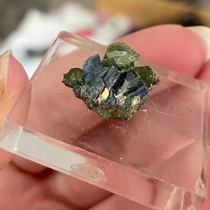 May include: A small, natural mineral specimen featuring a cluster of green and blue crystals. The crystals are set on a dark, textured base and encased in a clear, rectangular acrylic block. The specimen is held in a person's hand.
