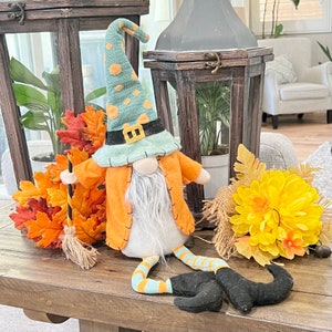 May include: A whimsical fall gnome with an orange coat, a mint green hat with orange polka dots, and black boots. The gnome has a long white beard and is holding a broom. Decorative fall leaves and a yellow flower arrangement are also present.