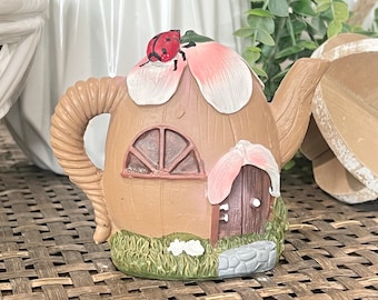 May include: A whimsical, brown ceramic teapot-shaped fairy house with a ladybug on top. The house features a door, window, and a spout. The roof is made of pink and white petals, and the base has green grass and a stone path.