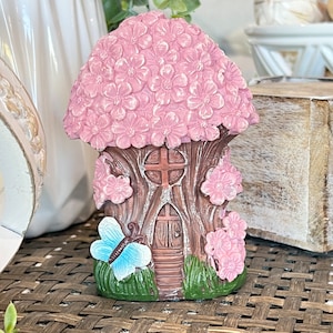 May include: A pink and brown fairy house with a butterfly and flowers. The house is made of resin and has a whimsical design.