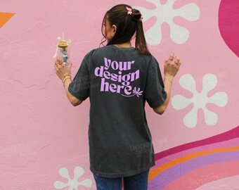 May include: A dark grey t-shirt with the text "your design here" in pink lettering. The shirt is being worn by a person standing in front of a pink wall with white flowers.