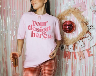 May include: A pink t-shirt with the text "your design here" in pink and red letters. The shirt is being worn by a person in a red and white checkered pattern pants. The background is a pink and white striped wall with a wreath and other decorations.