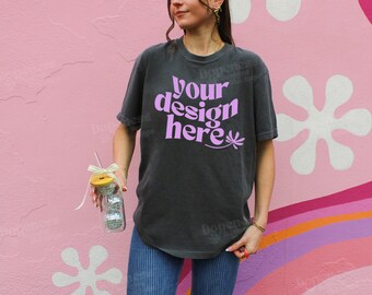 May include: A dark grey t-shirt with the text "your design here" in pink lettering. The shirt is being worn by a person in front of a pink and white wall with a rainbow pattern.