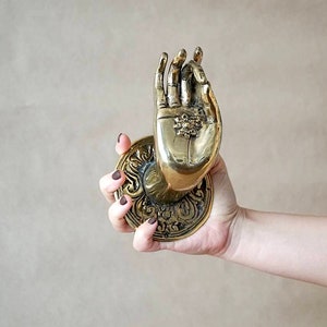 May include: A brass door knocker in the shape of a hand with a decorative floral design. The hand is in a welcoming gesture, with the palm facing up.